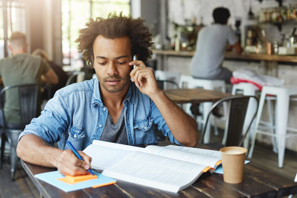 Homem estudando em um café enquanto faz anotações e fala ao telefone.