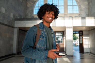 Jovem sorrindo em um hall de entrada iluminado, usando mochila e jaqueta jeans, segurando um smartphone enquanto olha para a câmera.