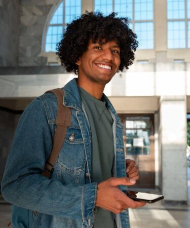Jovem sorrindo em um hall de entrada iluminado, usando mochila e jaqueta jeans, segurando um smartphone enquanto olha para a câmera.