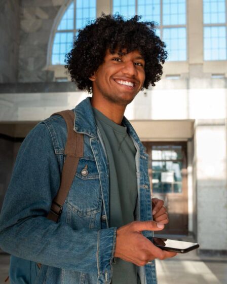 Jovem sorrindo em um hall de entrada iluminado, usando mochila e jaqueta jeans, segurando um smartphone enquanto olha para a câmera.