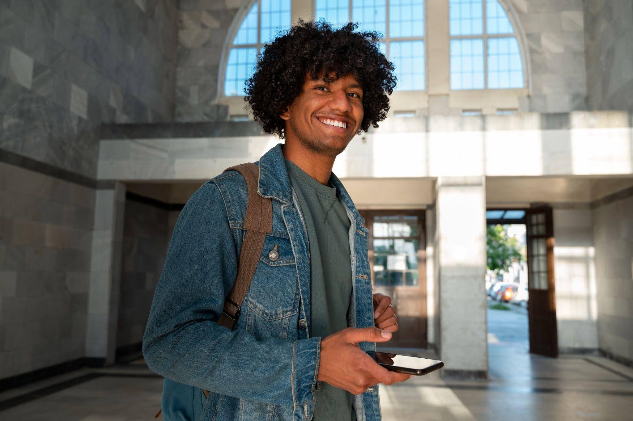 Jovem sorrindo em um hall de entrada iluminado, usando mochila e jaqueta jeans, segurando um smartphone enquanto olha para a câmera.