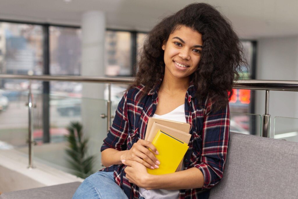 Mulher sorrindo sentada em um sofá, segurando livros e um caderno amarelo, em um ambiente interno moderno e iluminado por luz natural.