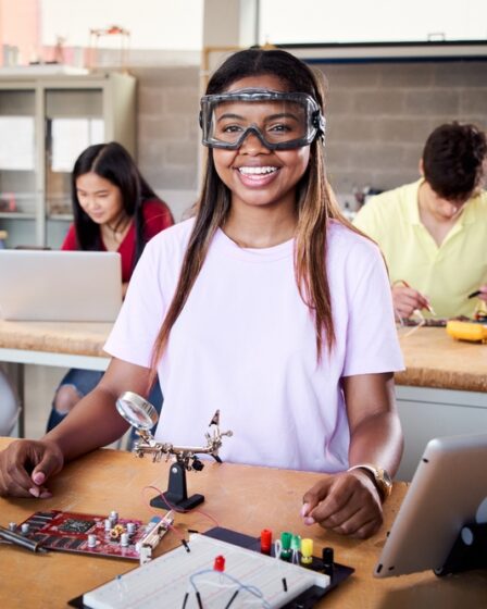 Menina sorri e olha para a câmera em um cenário com estudantes que mexem e estudam componentes eletrônicos.