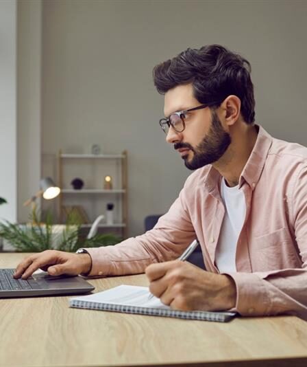 Homem mexe em seu laptop enquanto faz anotações em um caderno.
