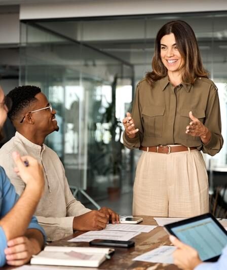 Uma mulher com semblante feliz em um escritório em conversa leve com sua equipe composta por dois homens e uma outra mulher, também com semblantes felizes.