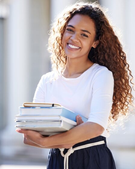 Jovem estudante negra, com cabelos longos cacheados, sorri enquanto segura livros e cadernos.
