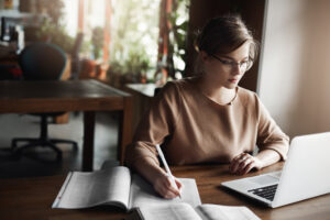 Uma mulher estudando muito concentrada em uma mesa com um caderno de anotações, um livro e um notebook.