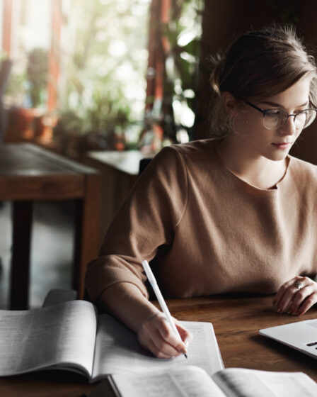 Uma mulher estudando muito concentrada em uma mesa com um caderno de anotações, um livro e um notebook.