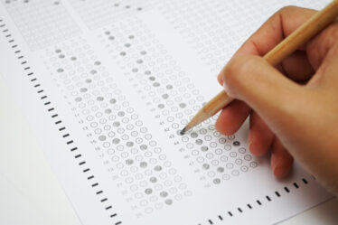 Mão segurando uma caneta esferográfica preta assinalando questões de uma prova em um caderno de respostas.