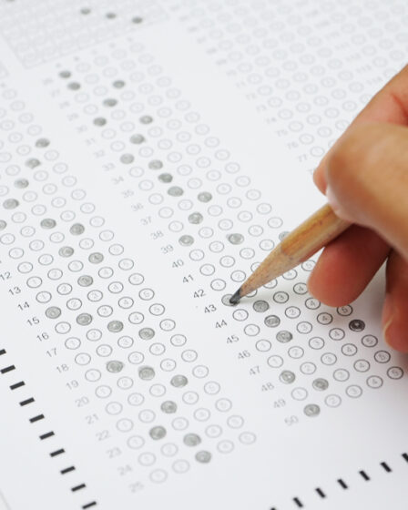 Mão segurando uma caneta esferográfica preta assinalando questões de uma prova em um caderno de respostas.