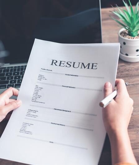 Pessoa segura currículo e uma caneta em cima de uma mesa com um laptop.