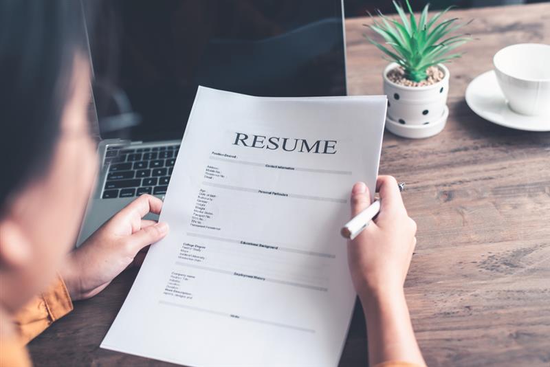 Pessoa segura currículo e uma caneta em cima de uma mesa com um laptop.