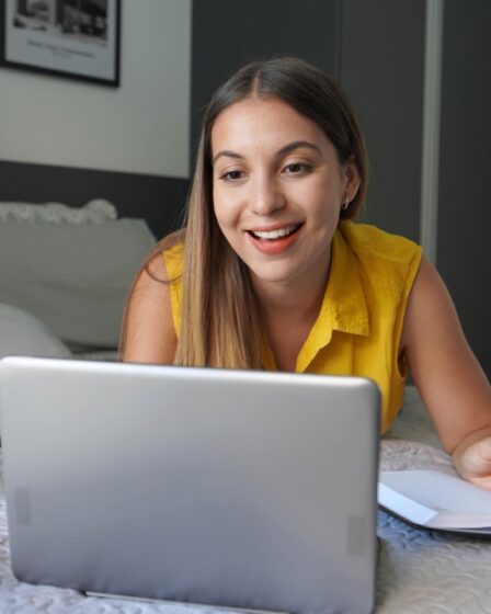 Uma mulher estudando com um semblante interessado e animado em uma cama com um caderno de anotações e um notebook.