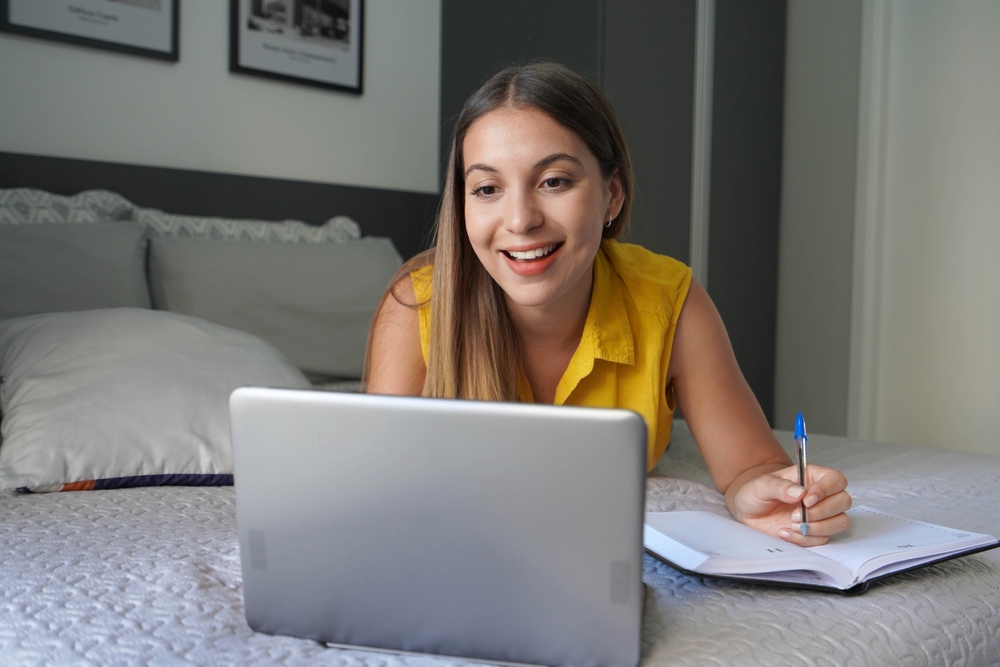 Uma mulher estudando com um semblante interessado e animado em uma cama com um caderno de anotações e um notebook.
