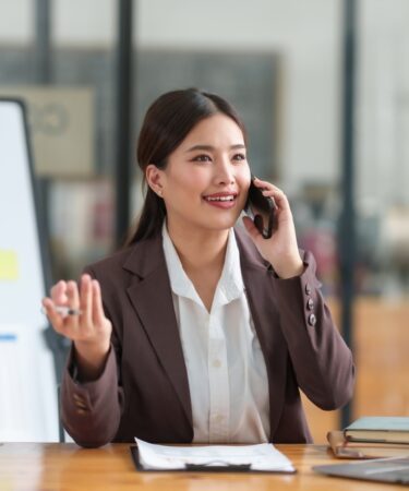 Uma mulher falando ao telefone ao lado de uma tela com gráficos sentada em uma cadeira em frente a uma mesa com uma prancheta e um notebook.