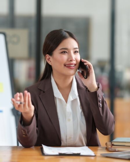 Uma mulher falando ao telefone ao lado de uma tela com gráficos sentada em uma cadeira em frente a uma mesa com uma prancheta e um notebook.
