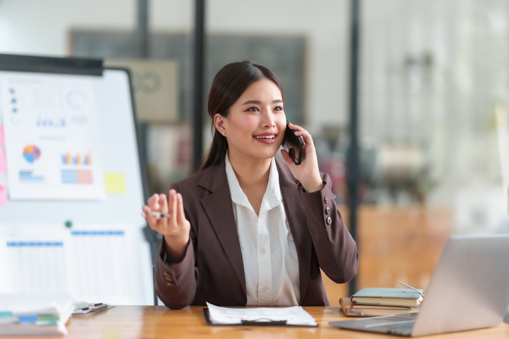 Uma mulher falando ao telefone ao lado de uma tela com gráficos sentada em uma cadeira em frente a uma mesa com uma prancheta e um notebook.