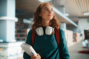 Mulher jovem usando um headfone olha para cima rindo enquanto segura um livro.