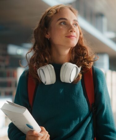 Mulher jovem usando um headfone olha para cima rindo enquanto segura um livro.
