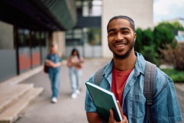 Jovem estudante universitário sorridente no campus da universidade, carregando cadernos e mochila, com prédios ao fundo.