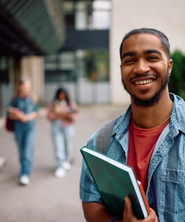 Jovem estudante universitário sorridente no campus da universidade, carregando cadernos e mochila, com prédios ao fundo.