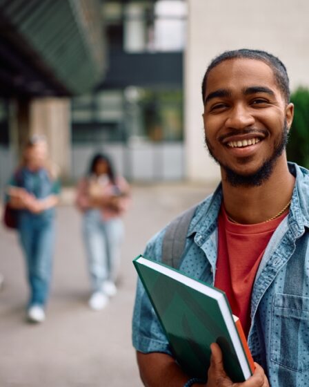Jovem estudante universitário sorridente no campus da universidade, carregando cadernos e mochila, com prédios ao fundo.