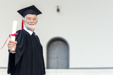 Idoso vestido com a beca segurando o diploma em direção a tela.