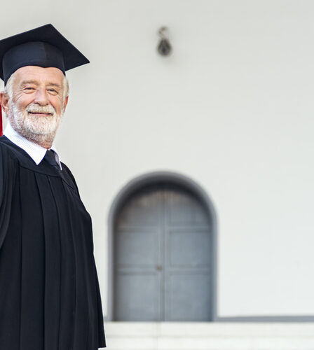 Idoso vestido com a beca segurando o diploma em direção a tela.