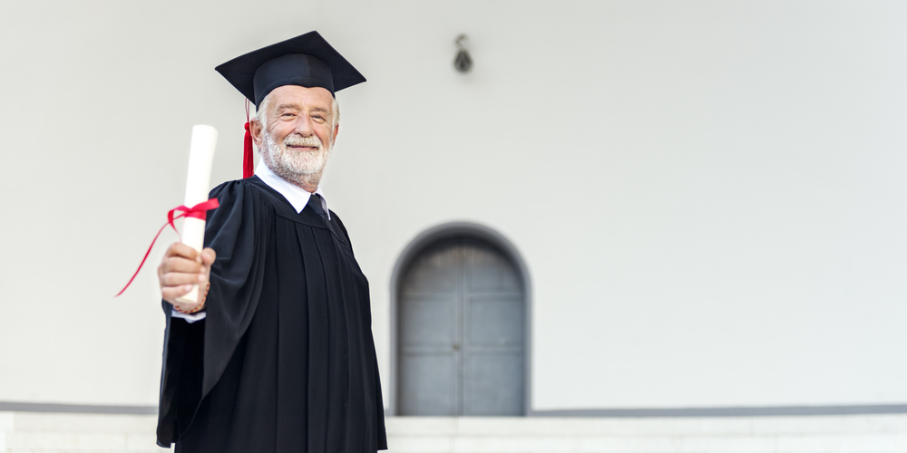 Idoso vestido com a beca segurando o diploma em direção a tela.