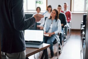 Professor palestrante em sala de aula com alunos atentos, simulando uma defesa de tese ou aula de doutorado e mestrado.
