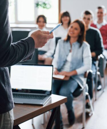 Professor palestrante em sala de aula com alunos atentos, simulando uma defesa de tese ou aula de doutorado e mestrado.