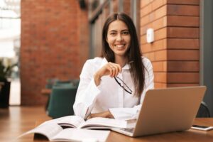 Mulher sorrindo com notebook e livros abertos na mesa, representando a rotina de pesquisas de cursos de doutorado e mestrado.