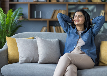 Mulher jovem, de cabelos longos e escuros, usando camisa jeans e calça bege, sorri enquanto relaxa sentada em um sofá, com as mãos apoiadas atrás da cabeça.