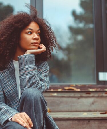 Mulher sentada em uma escada pensando apoiando o braço no joelho em pose pensativa.