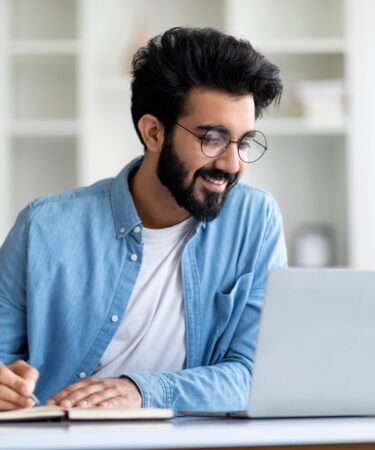 Homem jovem sorri em frente a um notebook, quanto faz anotações em caderno.