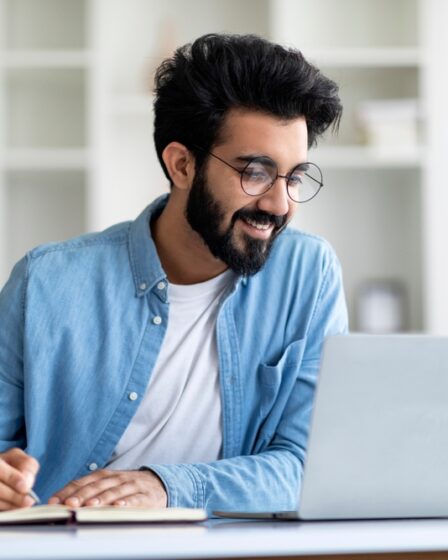 Homem jovem sorri em frente a um notebook, quanto faz anotações em caderno.