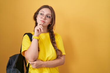 Jovem estudante com mochila olhando para o lado e expressando dúvida, em ambiente com parede amarelo de fundo.