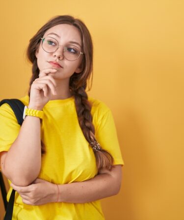 Jovem estudante com mochila olhando para o lado e expressando dúvida, em ambiente com parede amarelo de fundo.