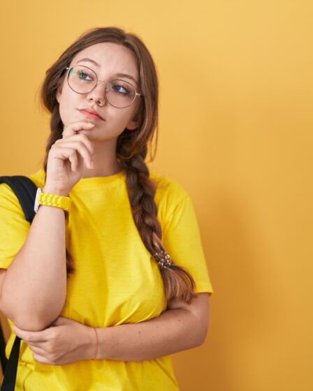 Jovem estudante com mochila olhando para o lado e expressando dúvida, em ambiente com parede amarelo de fundo.