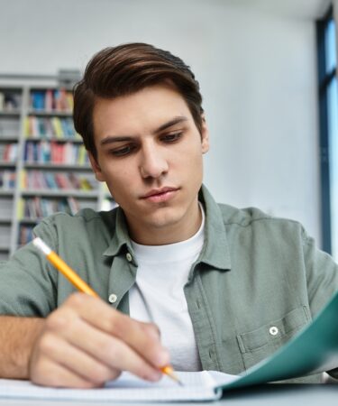Jovem estudante concentrado com a mão esquerda apoiando a capa de um caderno e a direita com o lápis na mão enquanto escreve nesse mesmo caderno.