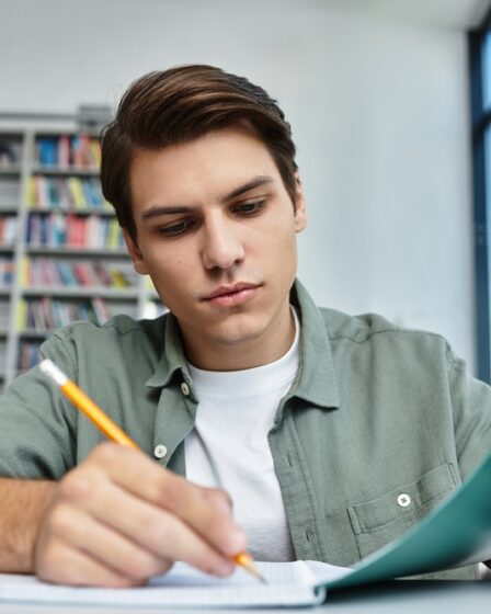 Jovem estudante concentrado com a mão esquerda apoiando a capa de um caderno e a direita com o lápis na mão enquanto escreve nesse mesmo caderno.