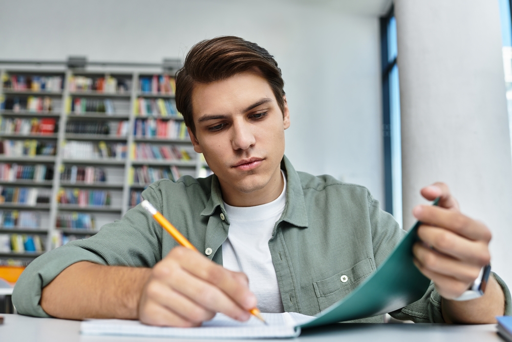 Jovem estudante concentrado com a mão esquerda apoiando a capa de um caderno e a direita com o lápis na mão enquanto escreve nesse mesmo caderno.