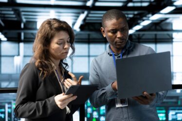 Dois profissionais, uma mulher e um homem, olham para um tablet e um notebook respectivamente.
