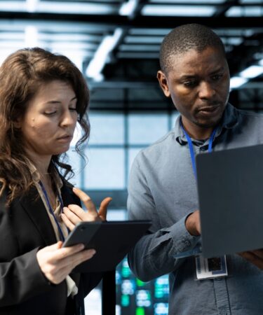Dois profissionais, uma mulher e um homem, olham para um tablet e um notebook respectivamente.