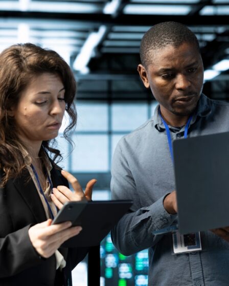 Dois profissionais, uma mulher e um homem, olham para um tablet e um notebook respectivamente.
