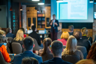 Imagem vista do fundo de uma plateia, onde homens e mulheres assistem a uma palestra.