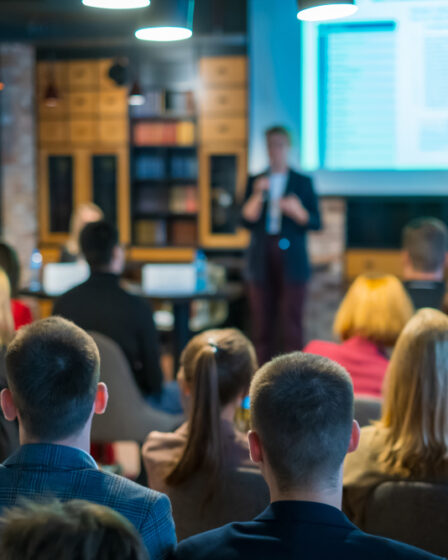 Imagem vista do fundo de uma plateia, onde homens e mulheres assistem a uma palestra.