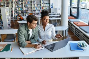 Um homem e uma mulher, jovens, estudando em uma biblioteca, sentados em uma carteira e utilizando um notebook e um caderno