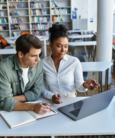 Um homem e uma mulher, jovens, estudando em uma biblioteca, sentados em uma carteira e utilizando um notebook e um caderno