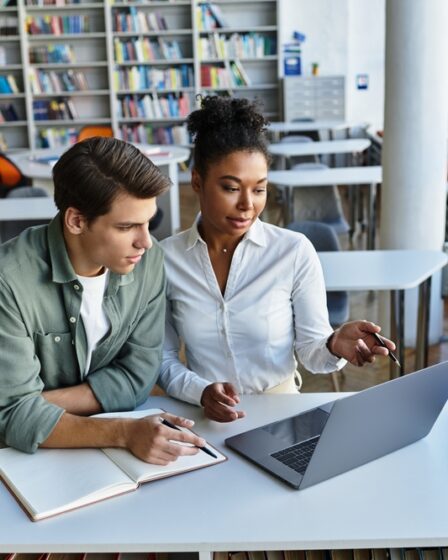Um homem e uma mulher, jovens, estudando em uma biblioteca, sentados em uma carteira e utilizando um notebook e um caderno
